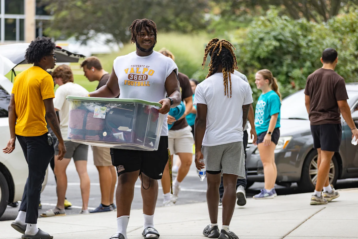 Smiling students carry storage bins and bags across the campus outdoors during college move-in day.