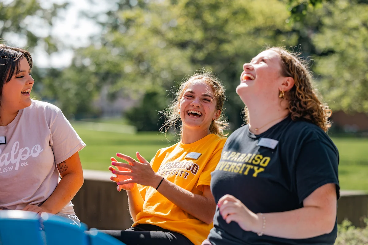 Three young women sit outside laughing together on a sunny day, wearing Valparaiso University shirts and chatting about their latest visit to the Registrar.