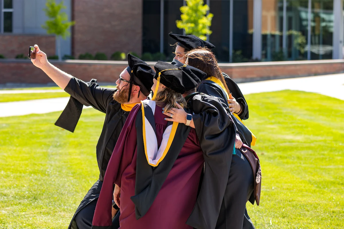 Four graduates in caps and gowns take a group selfie outside on a sunny day, celebrating their achievements after picking up their diplomas from the Registrar.