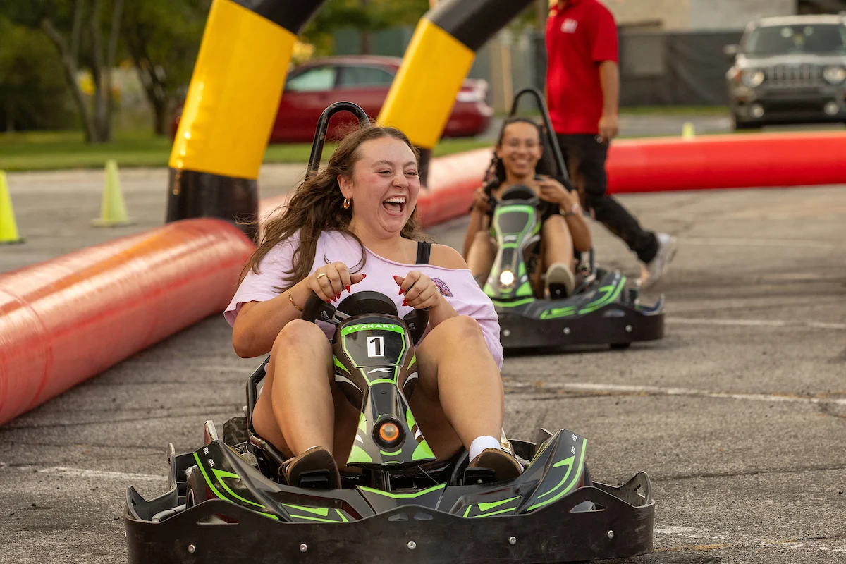 Two young women laughing and driving go-karts on an outdoor track, showcasing the joy of student involvement, with a person standing in the background.