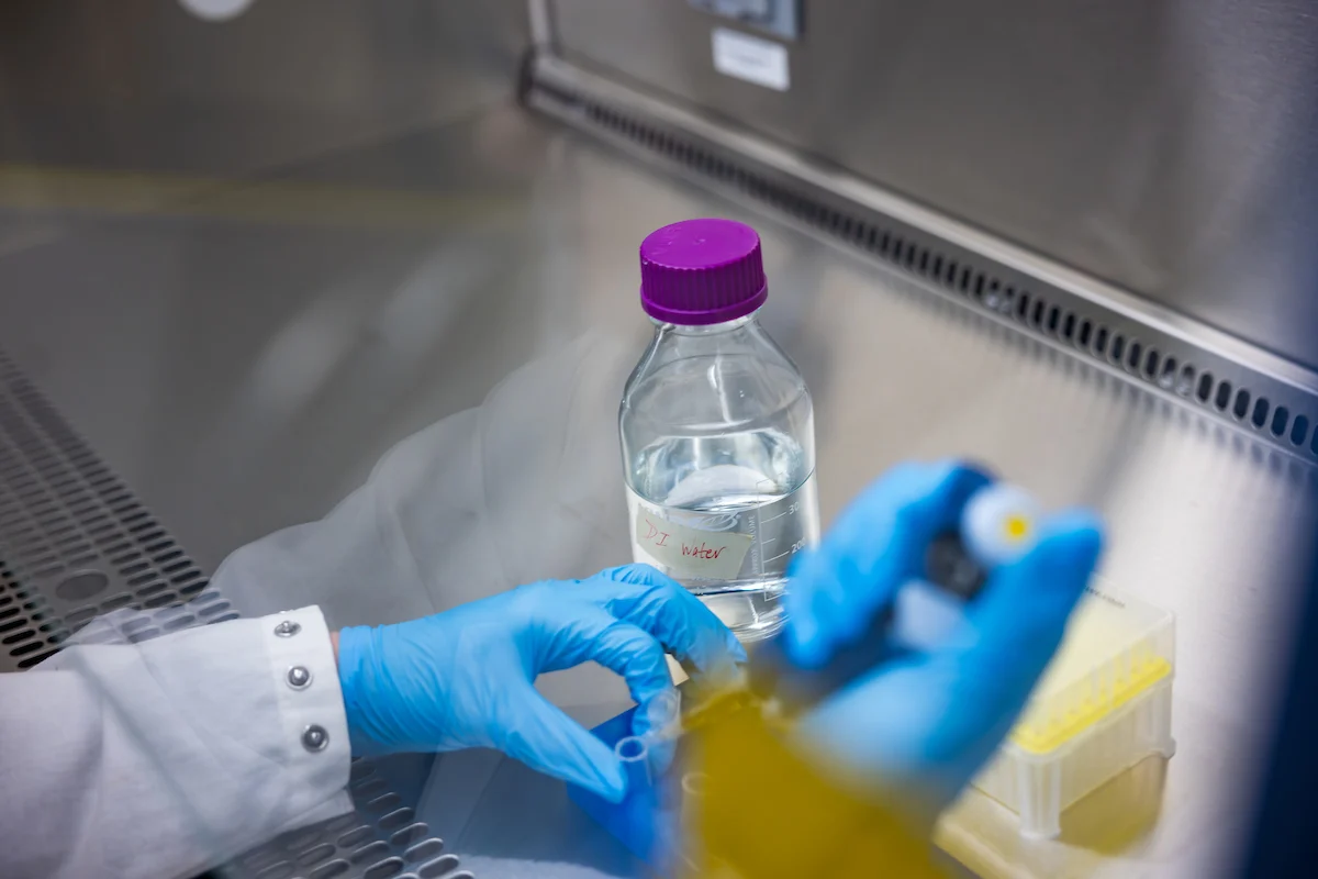 Gloved hands working with lab equipment near a clear bottle with a purple cap in a laboratory setting, demonstrating Campus Health and Safety protocols.