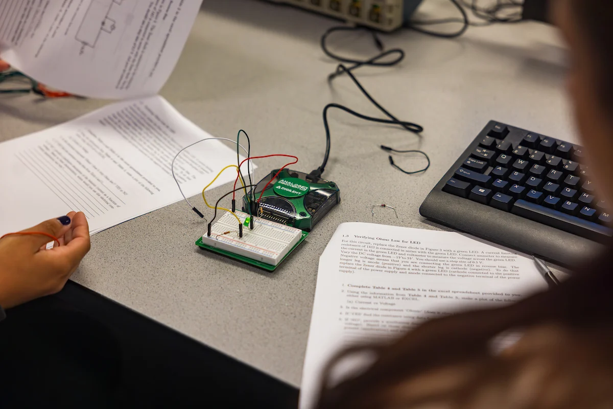 A person works on a breadboard circuit with wires next to printed instructions and a computer keyboard, capturing the hands-on learning atmosphere of campus life.