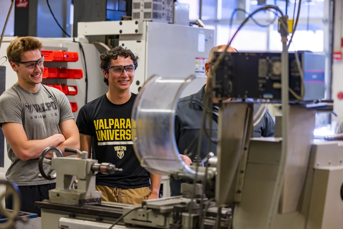 Three students wearing safety glasses smile while observing equipment in a university engineering workshop, demonstrating a strong commitment to Campus Health and Safety.