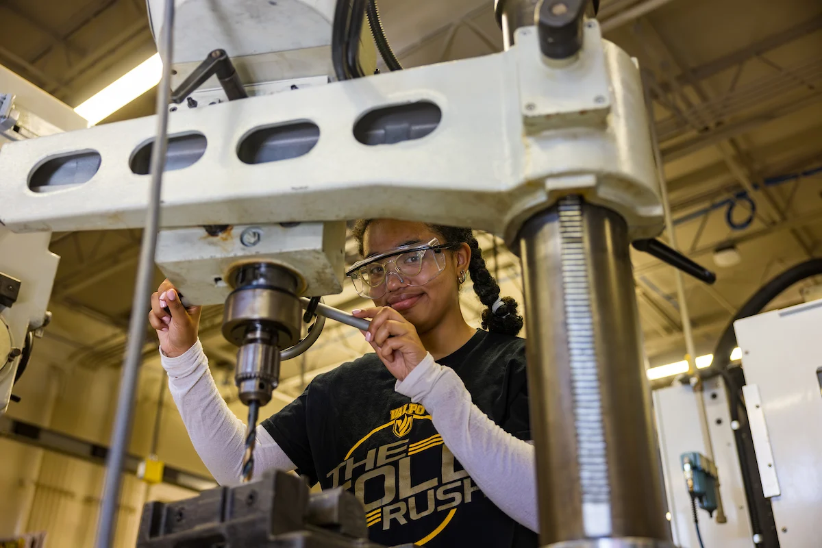 A person wearing safety glasses operates a large industrial drill press in a workshop, demonstrating the importance of Campus Health and Safety practices.