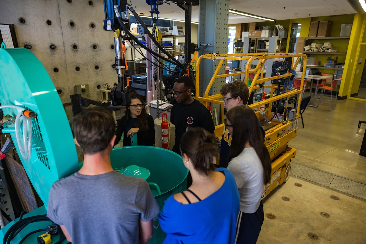 Six people gather around a large teal machine in an industrial lab setting, discussing or observing its function while prioritizing Campus Health and Safety.
