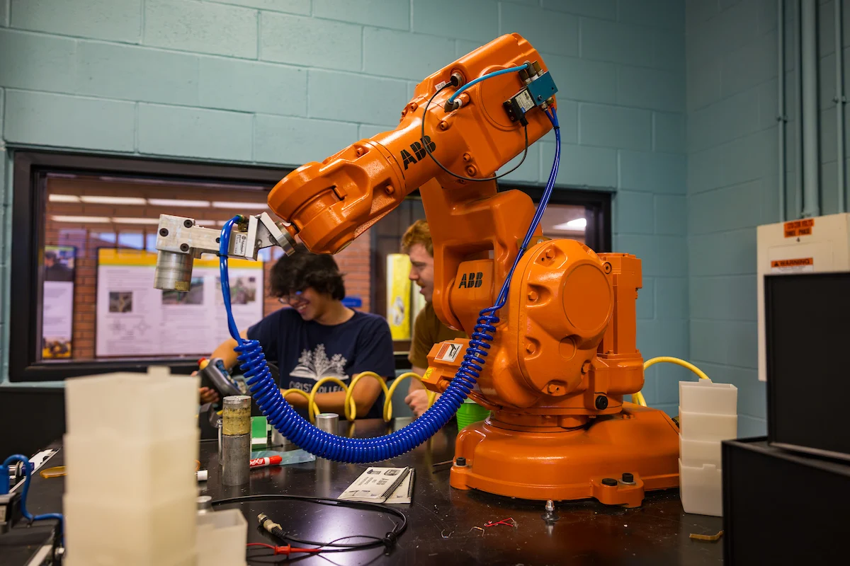 An orange industrial robotic arm operates in a lab with two people working in the background, adhering to Campus Health and Safety guidelines.