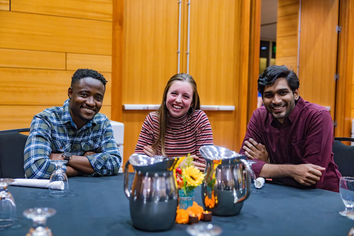 Three people smiling and sitting together at a table with pitchers, glasses, and a small flower arrangement, enjoying a moment of student involvement.
