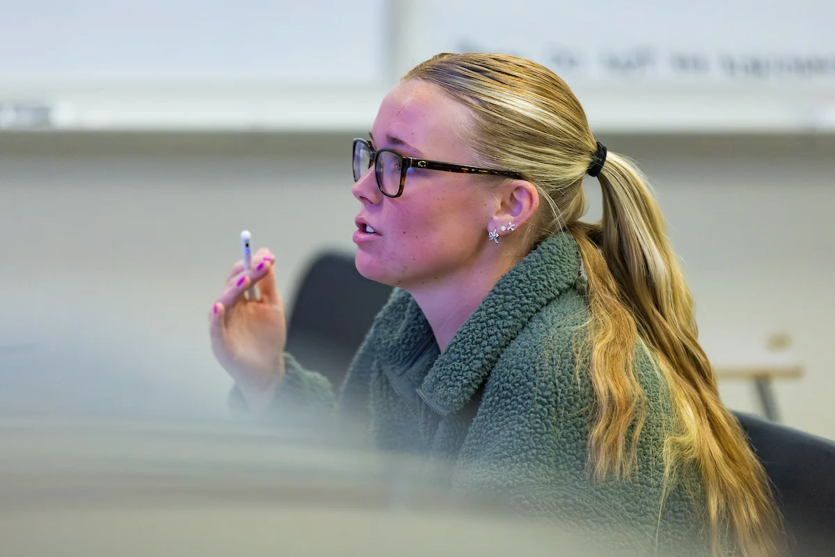 Young woman with glasses and a ponytail holds a pen and discusses transcript services in a classroom setting.