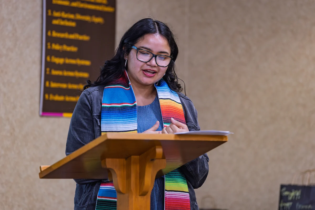 A woman wearing glasses speaks at a podium, wearing a colorful striped scarf, highlighting the importance of cultural diversity.