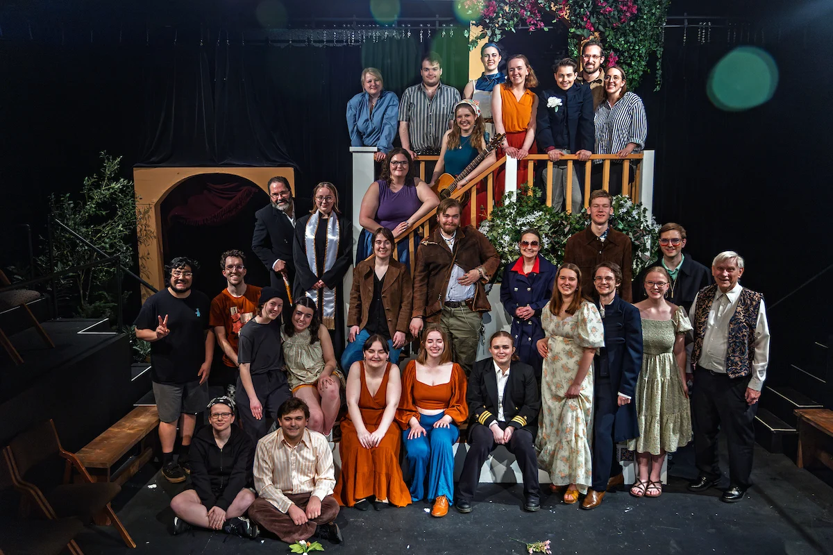 A theater cast in colorful costumes poses together onstage in front of a set with greenery and flowers, bringing campus life to the spotlight.