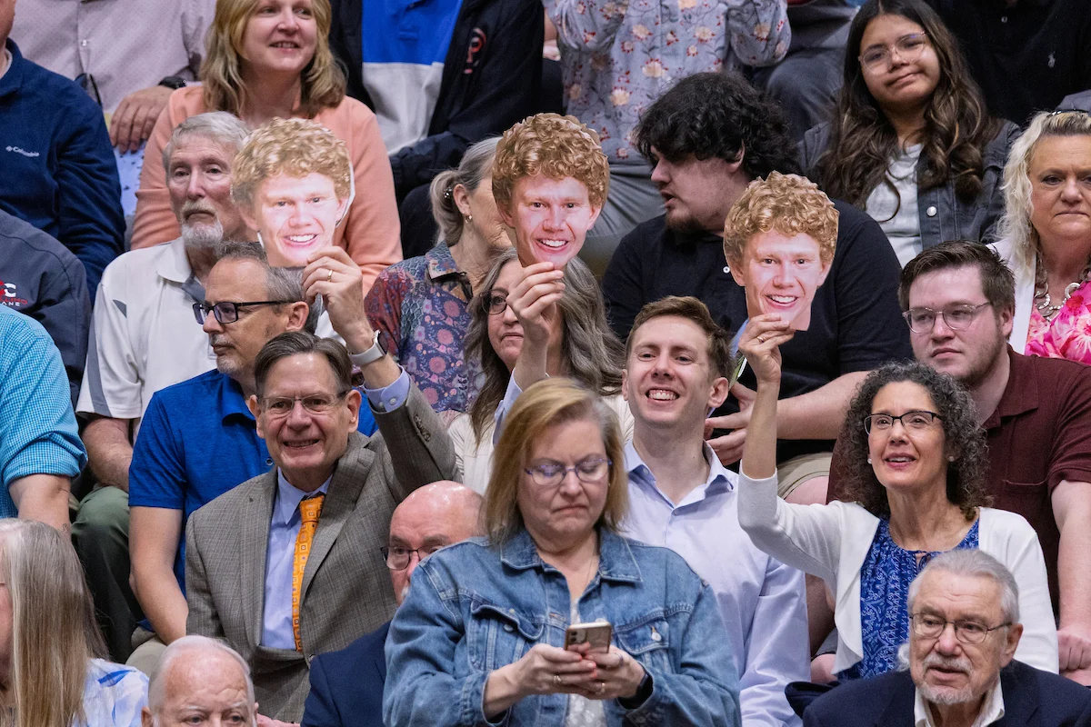 People in a crowd hold up face cutouts of a curly-haired man while others watch, use their phones, and search for Information for Graduates.