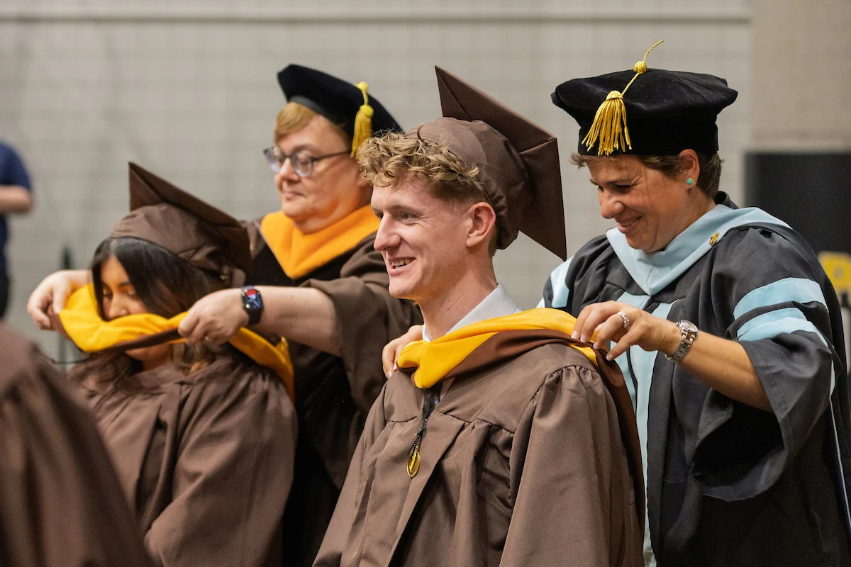 Graduates in brown gowns receive hoods during a commencement ceremony, smiling and celebrating their achievements.