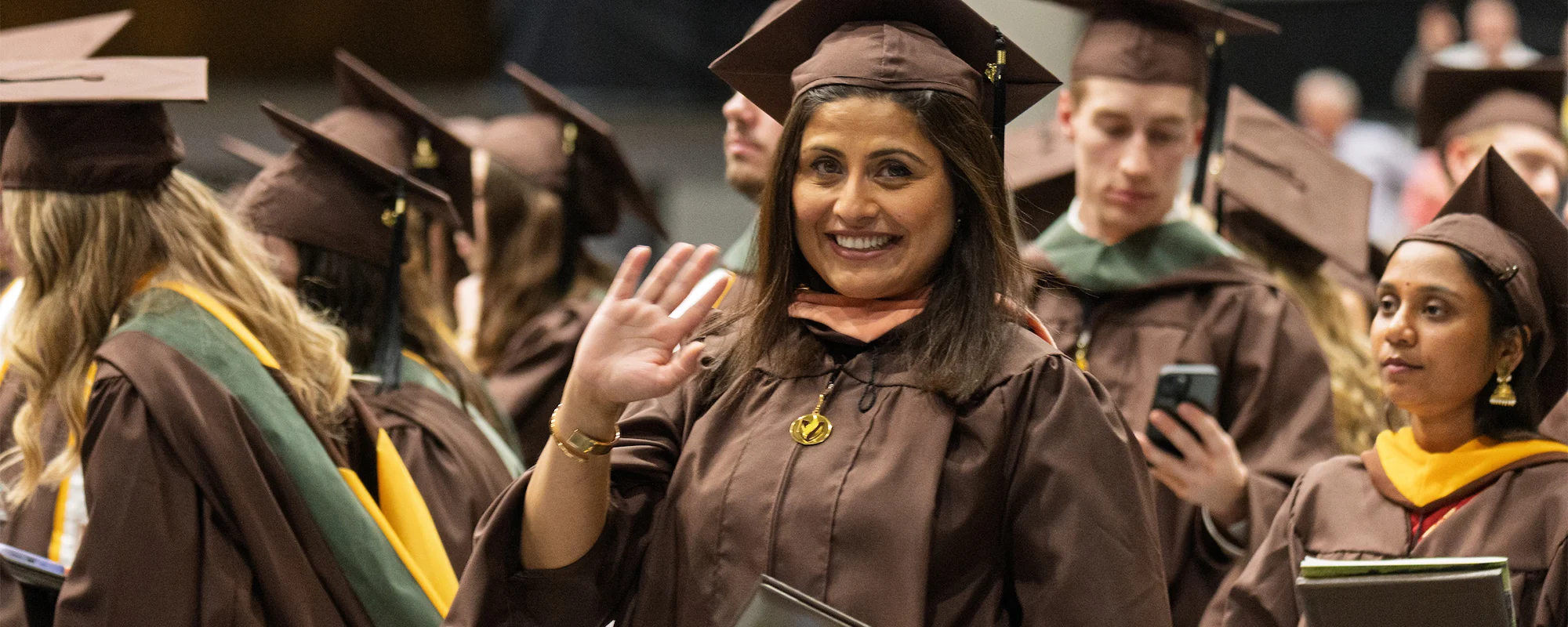Smiling graduate in cap and gown waves at the camera during a graduation ceremony.