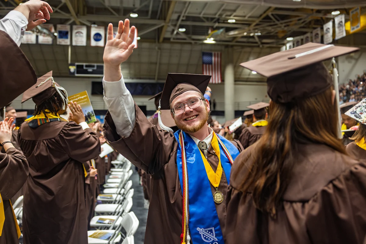 A smiling graduate in a cap and gown waves during a graduation ceremony in a large indoor arena, celebrating this milestone and sharing Information for Graduates.
