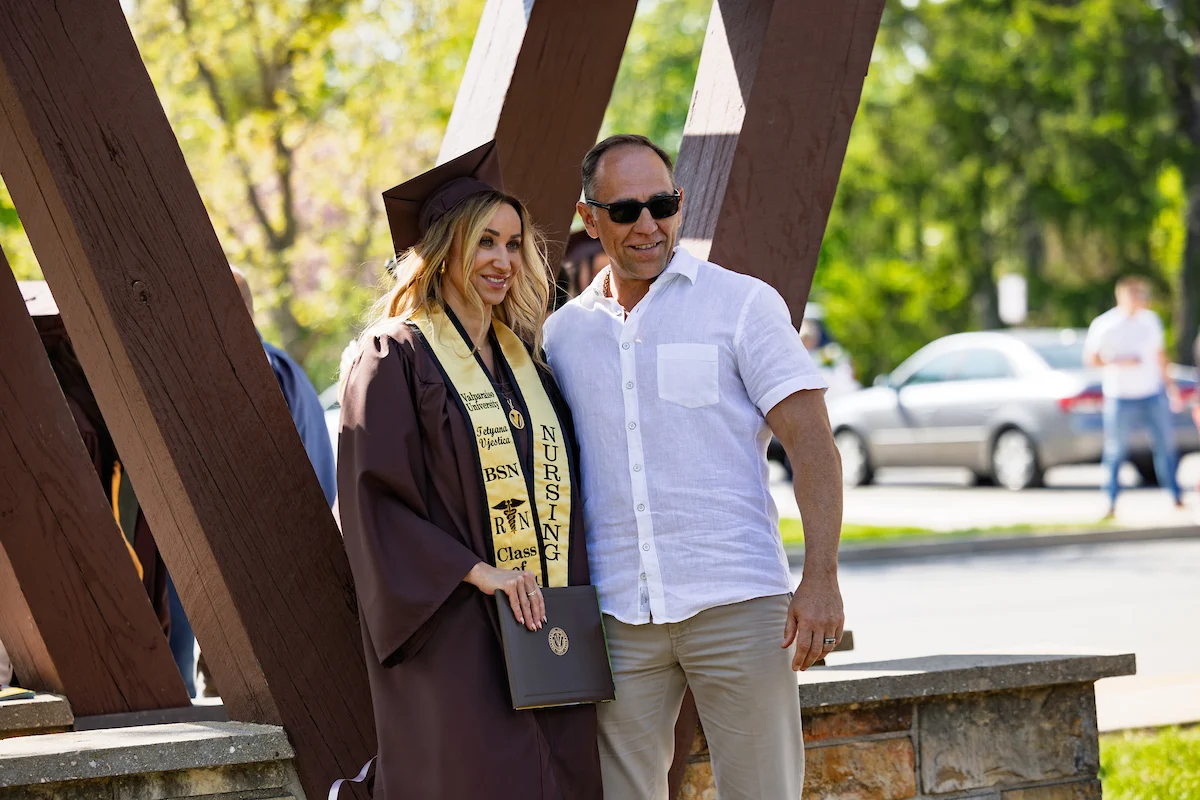 A woman in a graduation gown and sash stands smiling next to a man in sunglasses outdoors, ready to celebrate the big day—be sure to check Information for Guests for ceremony details.