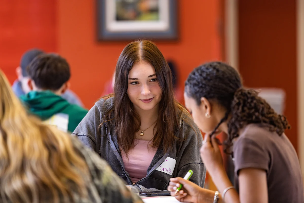 Two students sit at a table in discussion, one writing and the other listening attentively, as they review Registrar documents in a classroom setting.