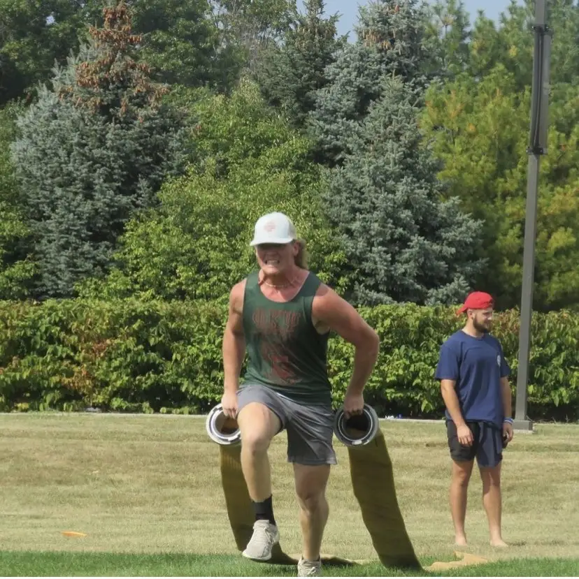 Abe Peetz carries rolled-up fire hoses on a grassy field, suggesting a moment of recreation, while another person stands in the background.