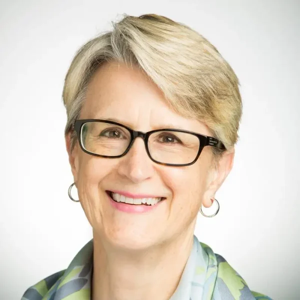 Smiling older woman with short blonde hair, glasses, and hoop earrings against a plain background—an inspiring figure in leadership education at Valpo LEAD.