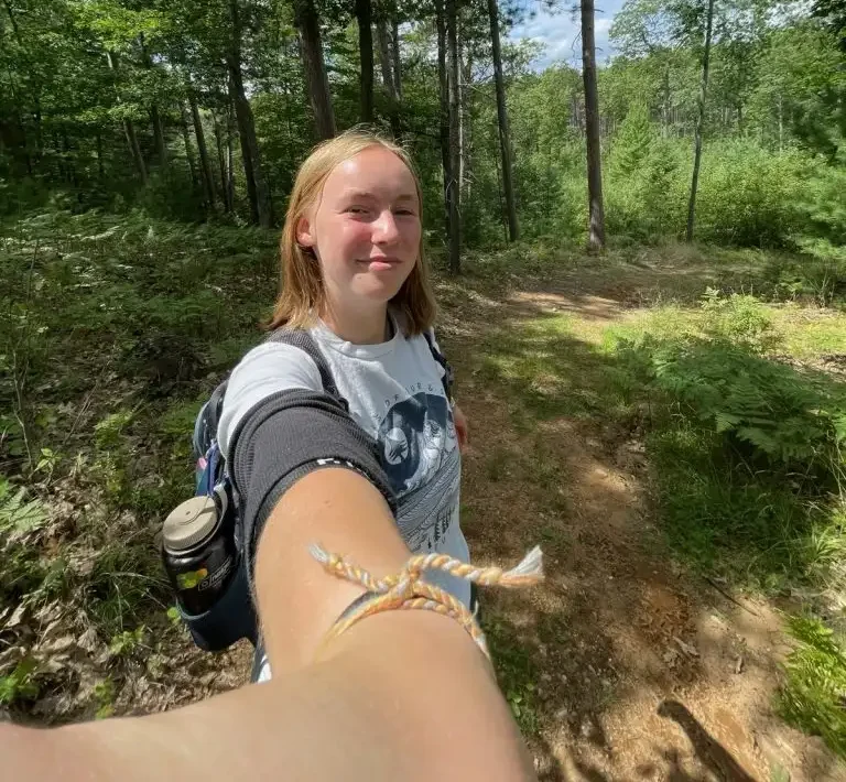 Emma takes a selfie while hiking on a forest trail, surrounded by tall green trees and sunlight, capturing her love for fitness and the great outdoors.