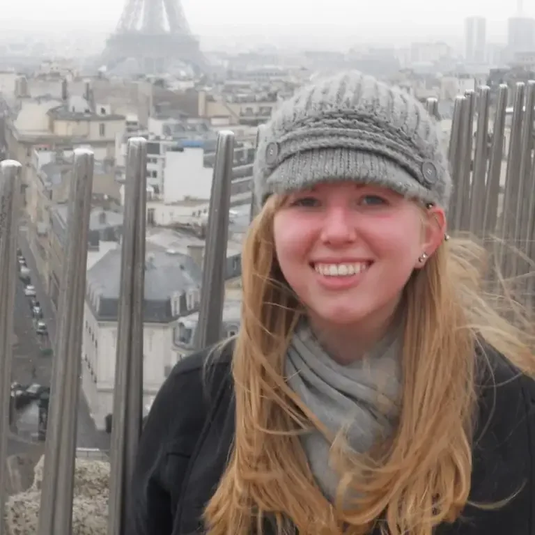 Smiling woman in a gray hat and scarf, celebrating her Placement Exam results with the Eiffel Tower and Paris cityscape in the background.