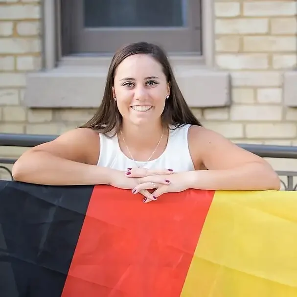 A woman smiles, leaning on a railing with a German flag draped in front of her, perhaps celebrating her success on a Placement Exam.