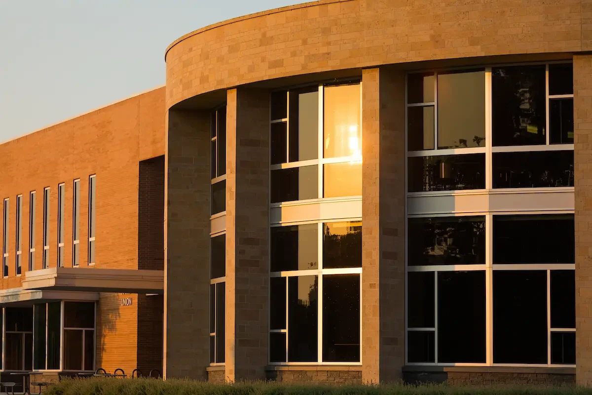 The sun reflects off large windows on the exterior of the modern brick Center for the Arts building at sunset.