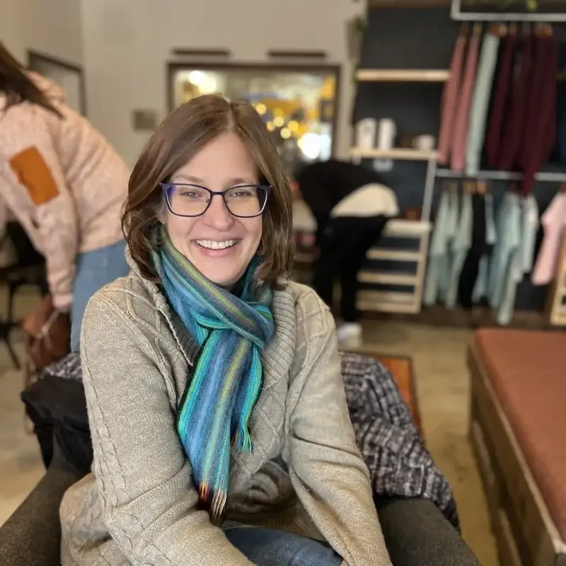Woman with glasses and a colorful scarf smiles while sitting indoors; clothes hang in the background, showing her support for LINC’s vibrant community.