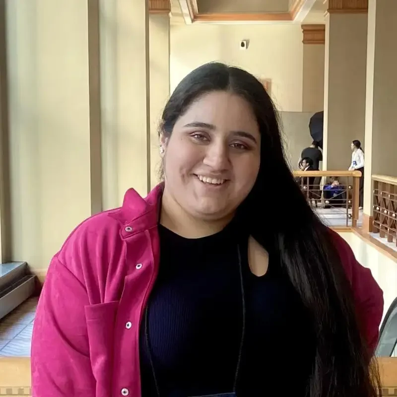 Person with long dark hair in a bright pink jacket smiling indoors by a sunlit hallway, ready to Extend Your Learning with positivity.
