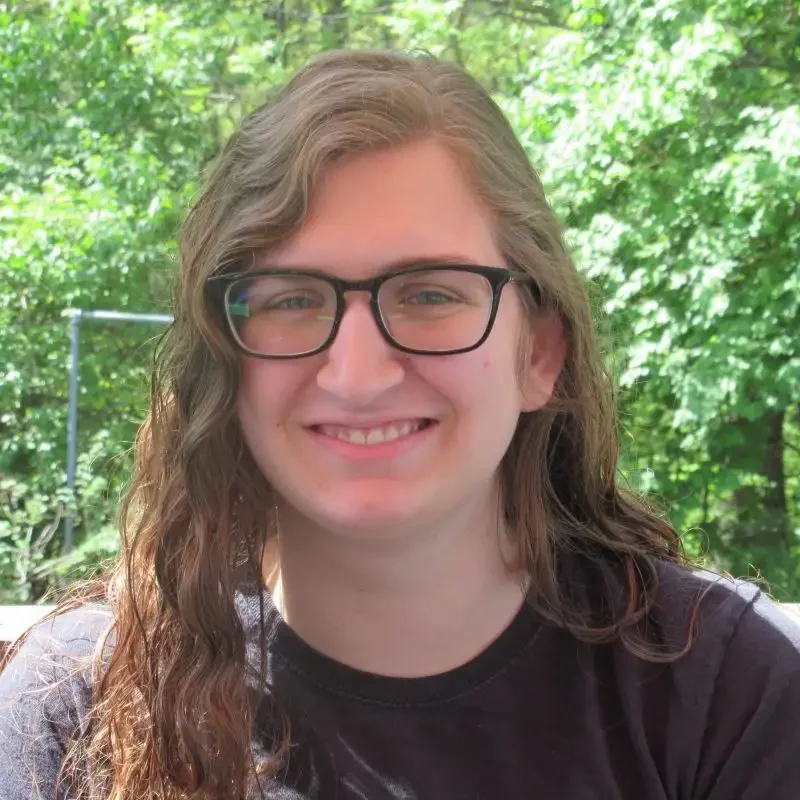 A person with long brown hair and glasses smiles outdoors, with green trees in the background—an ideal moment to Extend Your Learning amidst nature’s tranquility.