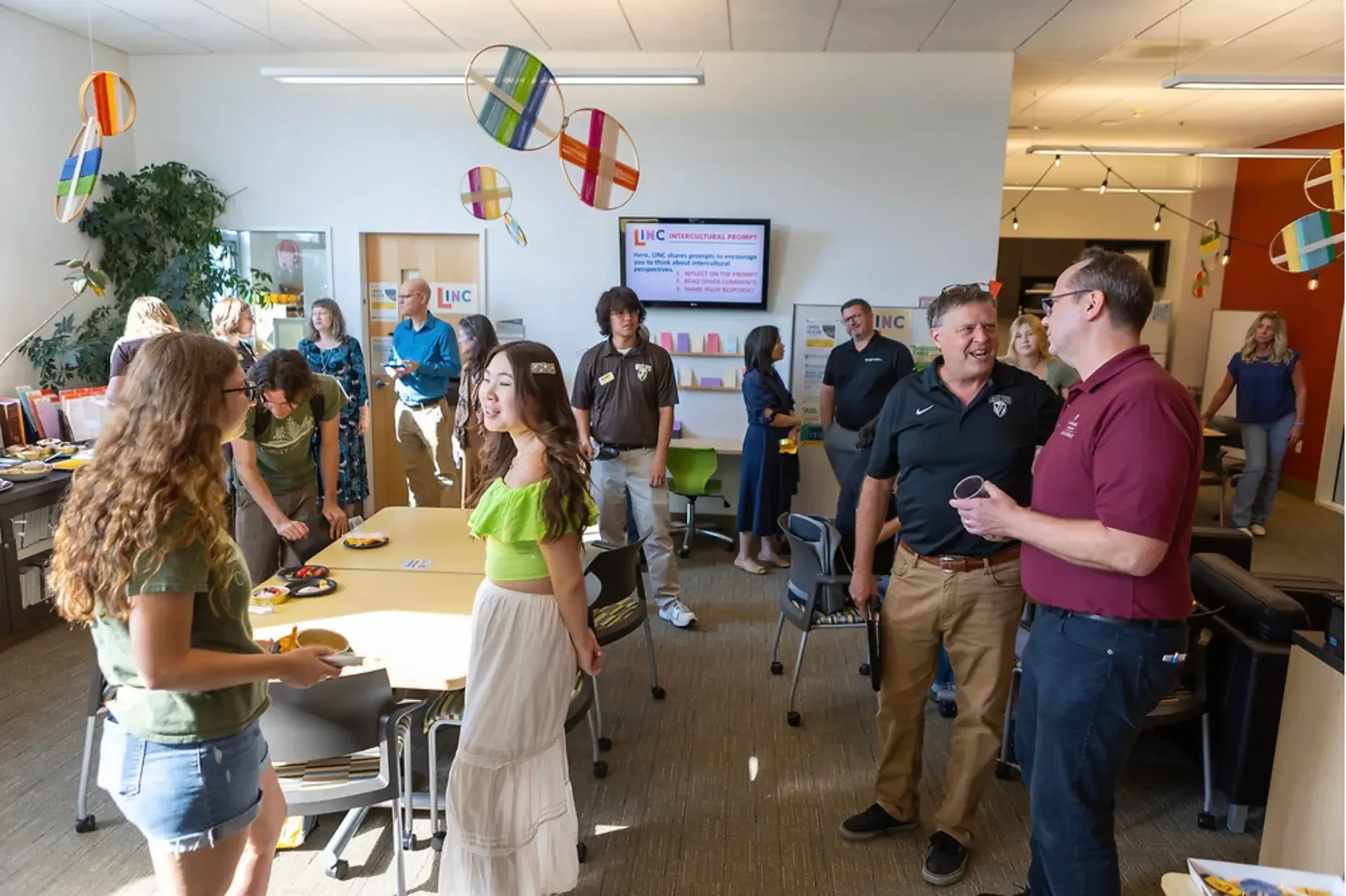 People gather and socialize in a brightly lit room adorned with colorful hanging ornaments, enjoying lively conversations.