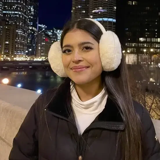 Woman wearing white earmuffs and a black coat smiles outside at night with city lights in the background, inspiring you to Extend Your Learning beyond the classroom and explore new experiences.