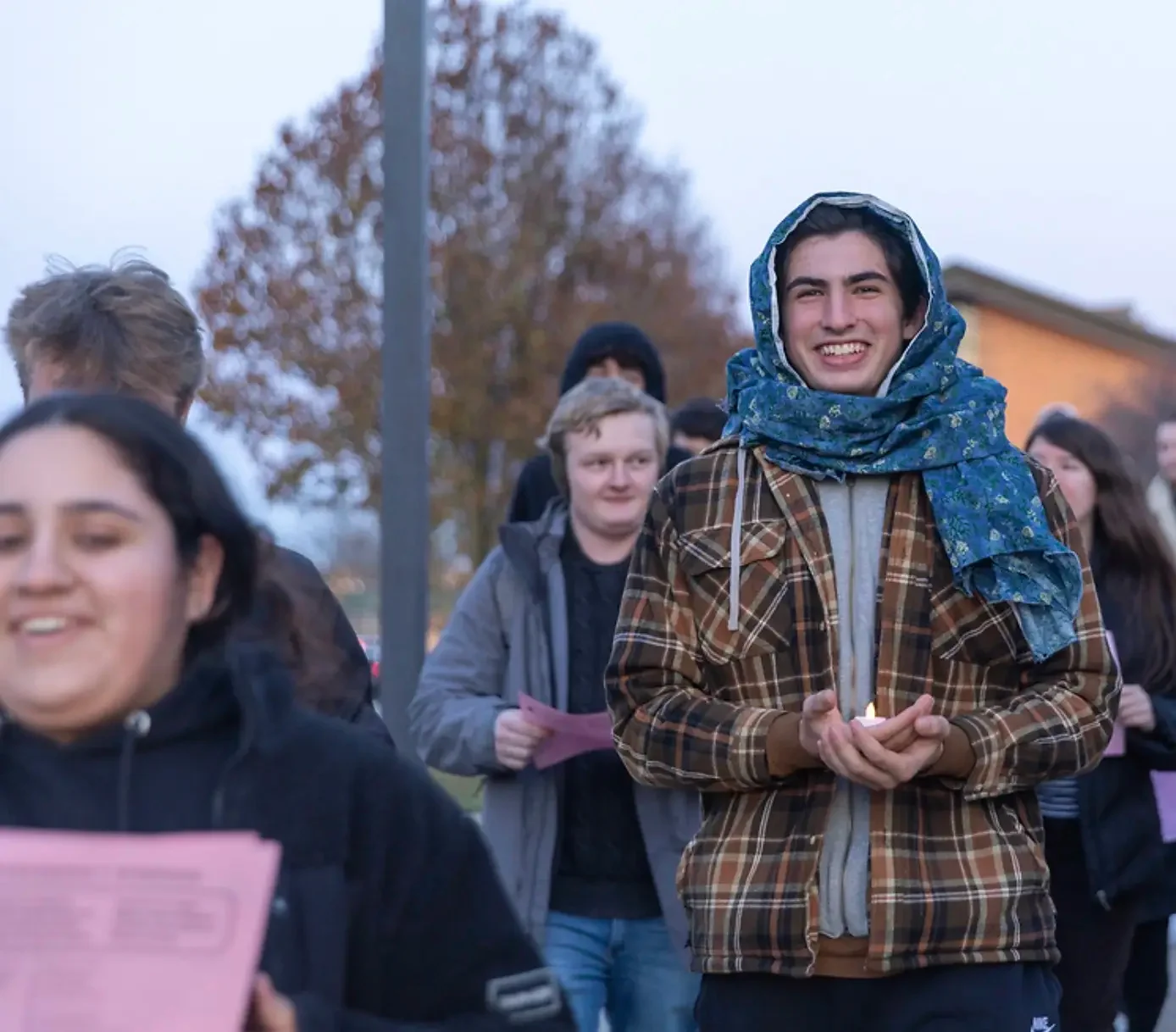 A group of people walking outside, one person smiling and wearing a blue scarf over their head stands out among the people.