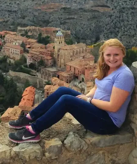 Woman in a purple shirt sits on a ledge overlooking a historic town with reddish buildings and a church, perhaps taking a break after her Placement Exam.