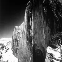 A steep, dramatic rock face rises above a snowy landscape under a clear sky, making it an inspiring subject for exhibitions & series exploring nature’s raw beauty.
