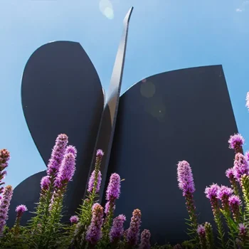 Large black butterfly sculpture behind purple flowers, set against a bright blue sky.
