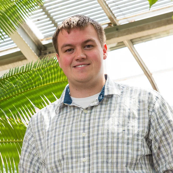 David Goad, in a plaid shirt, stands in a bright greenhouse surrounded by large green leaves.