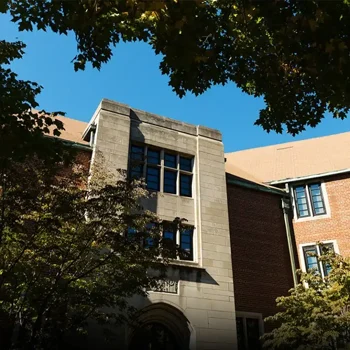 A brick and stone campus building framed by leafy trees under a clear blue sky.