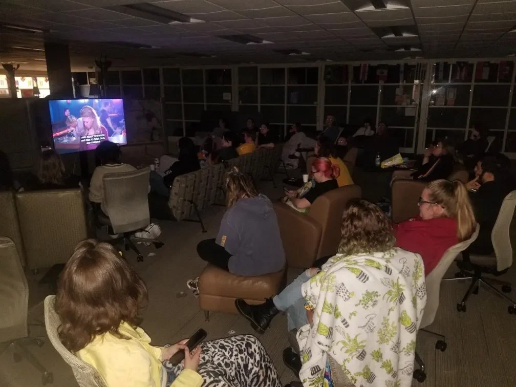 A group of people sit in a dim room watching a movie on a projector screen with subtitles.