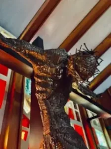 A metal sculpture of Jesus on the cross, featuring a crown of thorns, stands inside a historical chapel with exposed wooden beams.