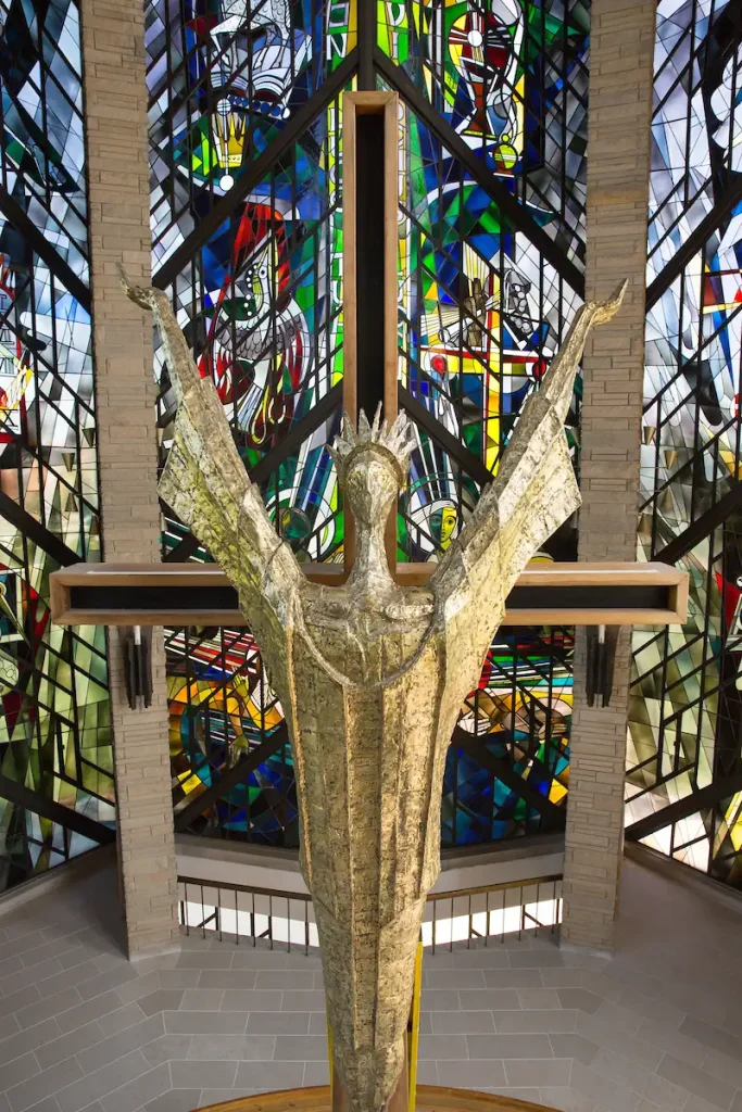 Golden statue of Christ with raised arms stands before colorful stained glass windows, celebrating chapel history.