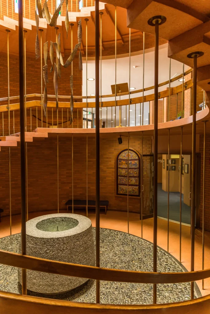 Spiral staircase and circular stone font reflect the History of the Chapel in a modern, brick-walled interior with metal railings.