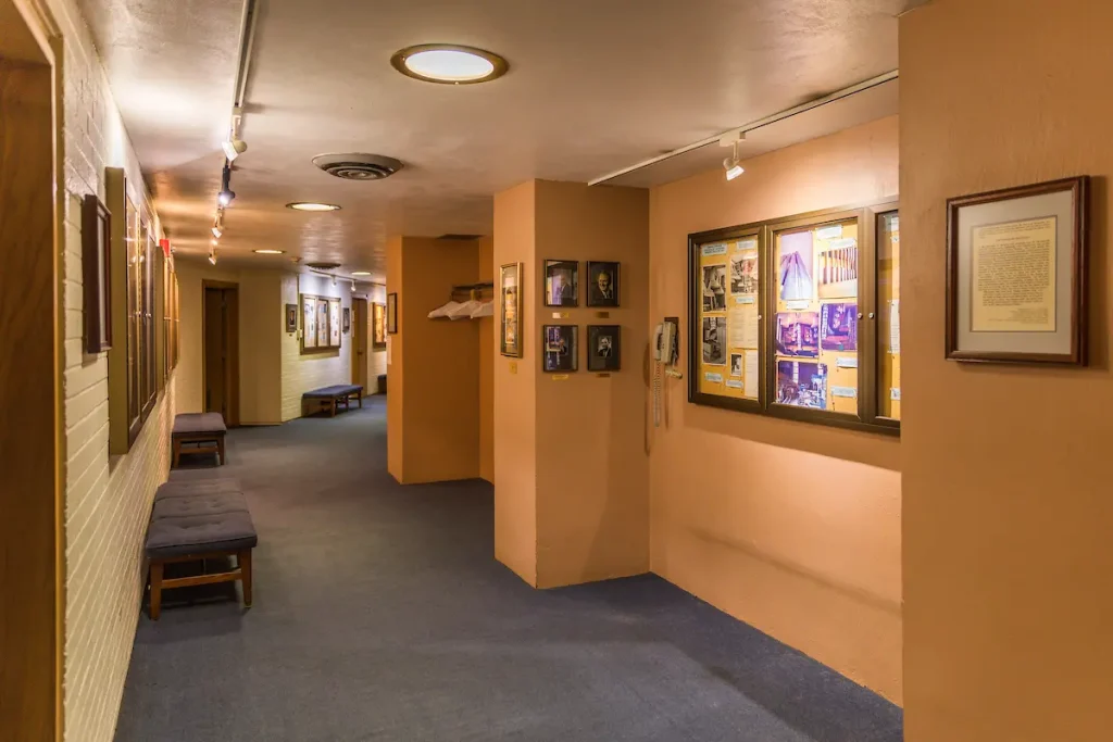 A hallway with framed photos and displays on peach-colored walls, blue carpet, and benches along the side highlights the rich history of the chapel.