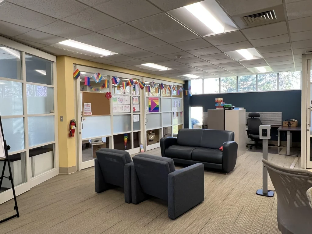 Office lounge with gray chairs, sofas, pride flags, and decorated glass walls, with desks and cabinets in back.