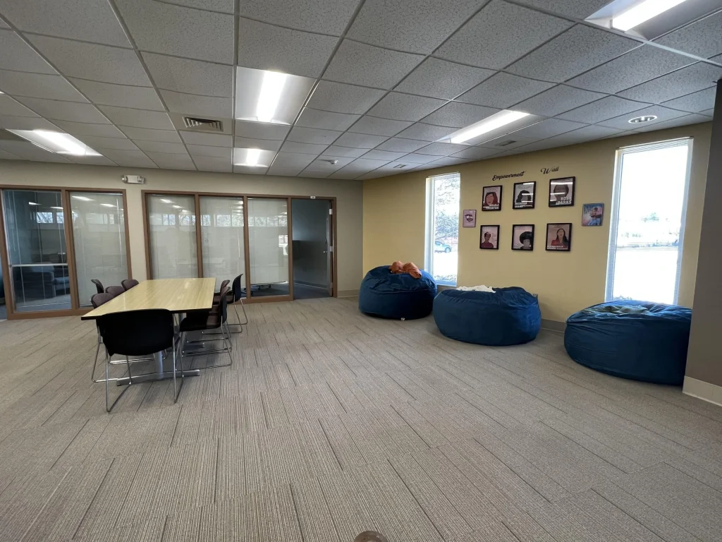 A modern lounge with bean bags, a long table with chairs, large windows, and framed photos on the wall.