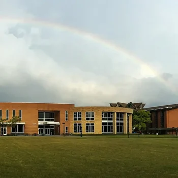 A faint rainbow arcs over a modern brick building and a green lawn under a cloudy sky.