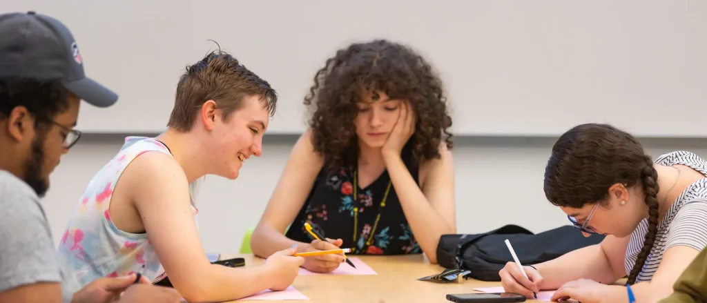 Four students sit at a table, writing and talking together in a classroom setting, sharing experiences as first-generation learners.