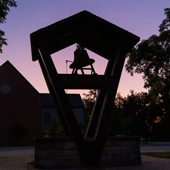 A bell in a wooden frame labeled "VICTORY" at sunset, silhouetted against a purple sky.