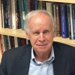 David Rowland, Ph.D., an older man in a suit, smiles warmly while sitting in front of a bookshelf filled with books.