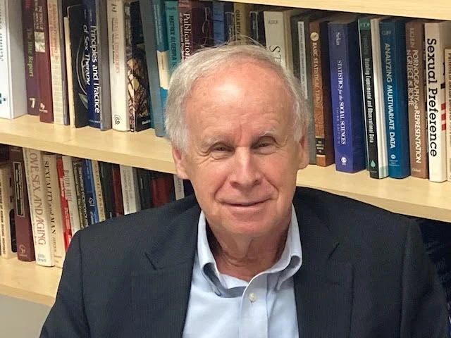 David Rowland, Ph.D., an older man in a suit, smiles warmly while sitting in front of a bookshelf filled with books.