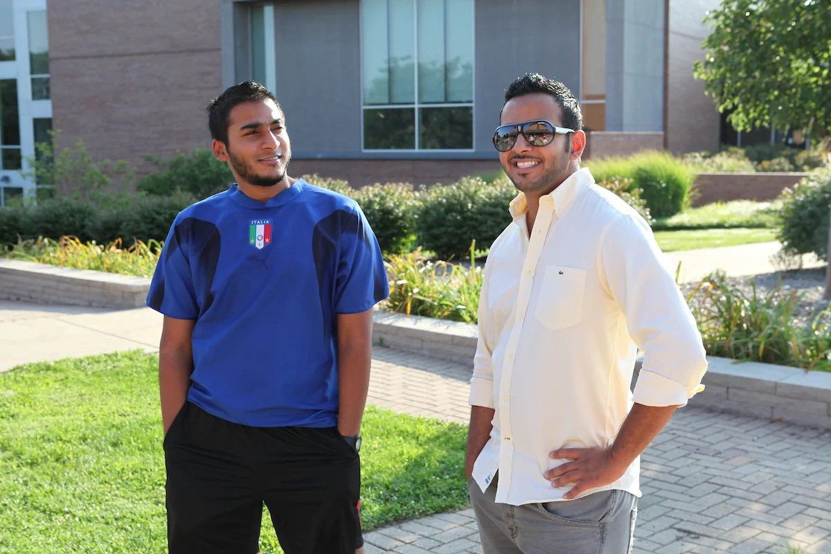 Two men standing outside, one in a blue shirt and the other in white, smiling and talking about international education on a sunny day.